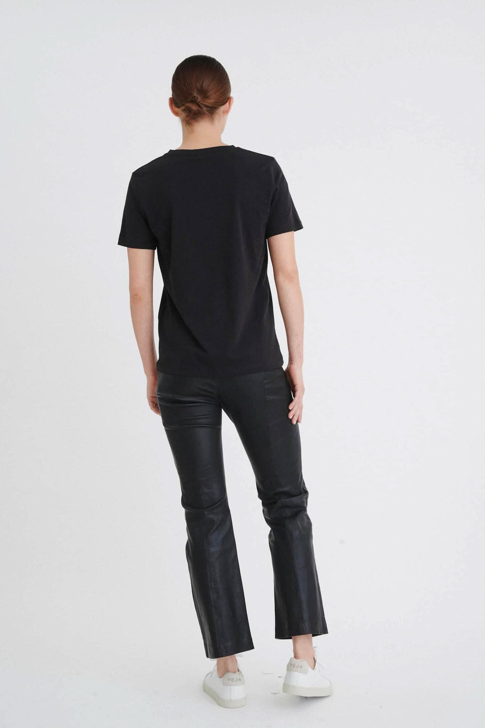 Femme portant un t-shirt noir col en V, vu de dos, avec un pantalon en cuir noir.