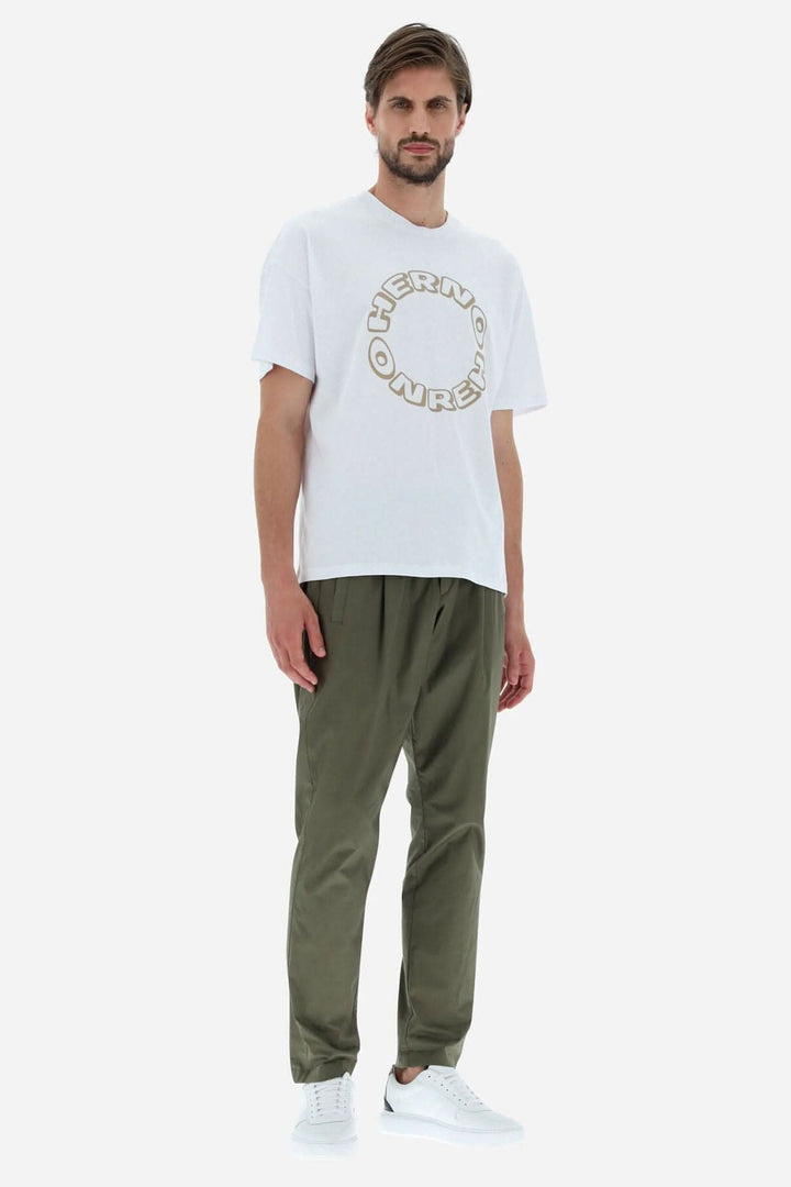 Homme portant un T-shirt blanc HERNO avec logo graphique, pantalon vert et baskets blanches, style décontracté.