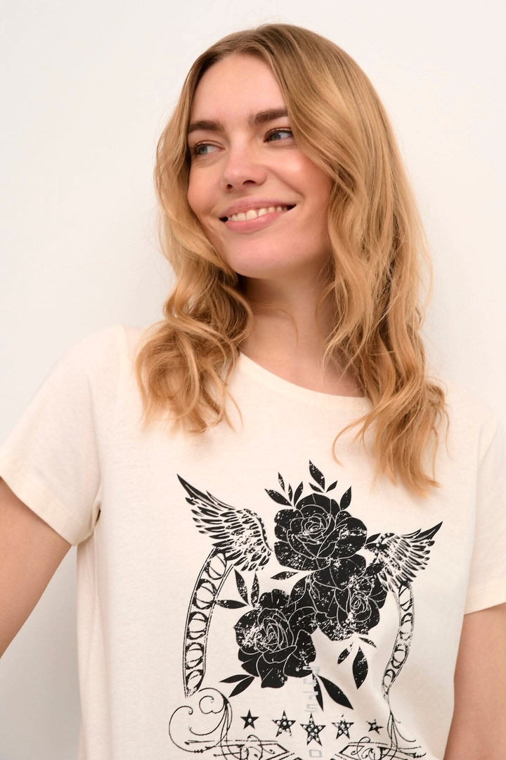 Femme souriante portant un t-shirt beige avec un imprimé floral noir sur le devant.