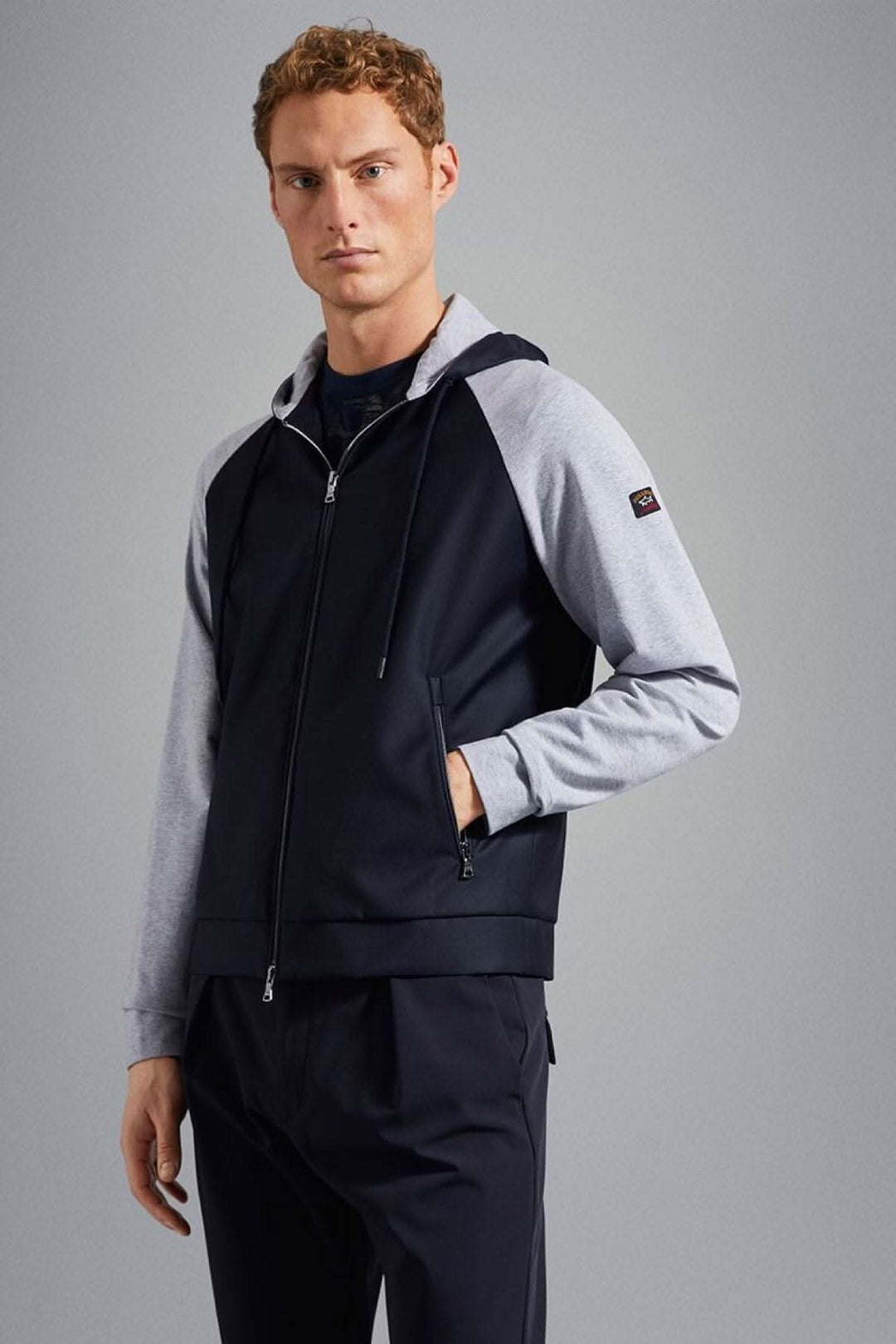 Homme portant une veste à capuche bleu marine avec logo, manches grises et poches latérales zippées.