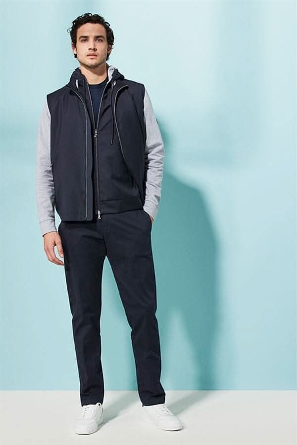 Homme portant une veste à capuche bleu marine avec logo, manches en tissus, pantalon noir et baskets blanches.